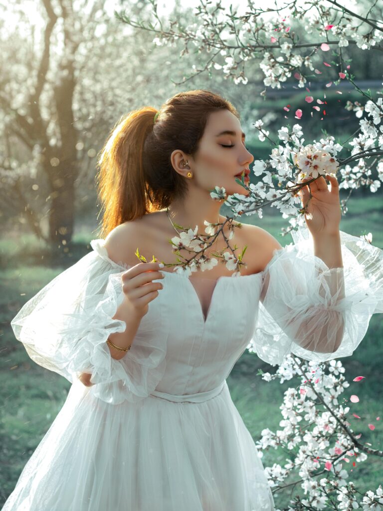 A bride in a flowing white dress admiring cherry blossoms outdoors.